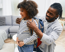 Father helps young daughter put on a backpack to prepare for school.