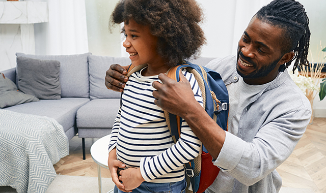 Father helps young daughter put on a backpack to prepare for school.