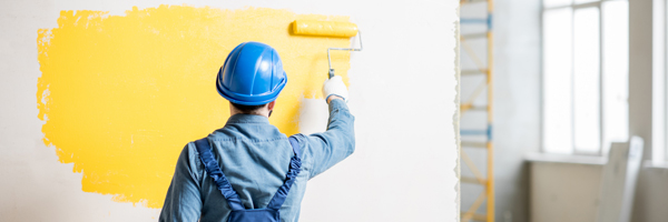 Man wearing a blue outfit paints a blank wall yellow with a roller brush.