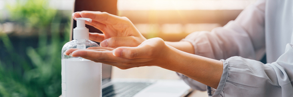 Woman uses hand sanitizer that sits on a wooden desk with a laptop.