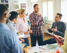 A group of six business employees sit and stand around a table talking during a meeting.