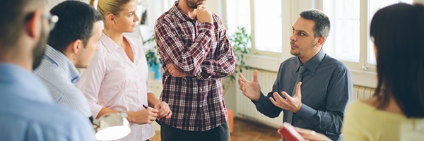 A group of six business employees sit and stand around a table talking during a meeting.