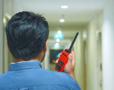 A man facing away from the camera holds a red radio up to his face and walks down an empty hallway.