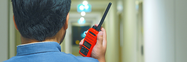 A man facing away from the camera holds a red radio up to his face and walks down an empty hallway.