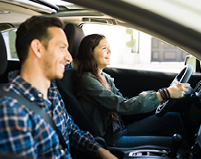 A teenage girl and her father smile and wear their seatbelts as they sit in the front of a car.