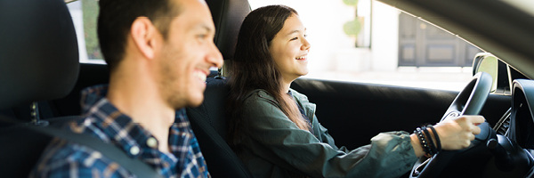 A teenage girl and her father smile and wear their seatbelts as they sit in the front of a car.