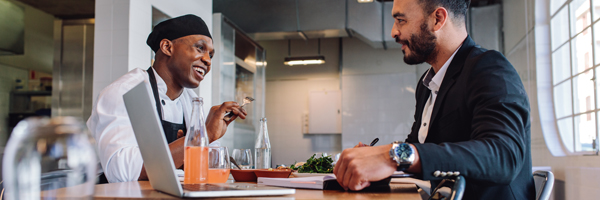 A man wearing an apron and a man wearing a business suit have a meeting in a restaurant.