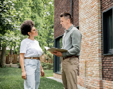 A realtor holds a folder and speaks to a woman outside a brick home in summer