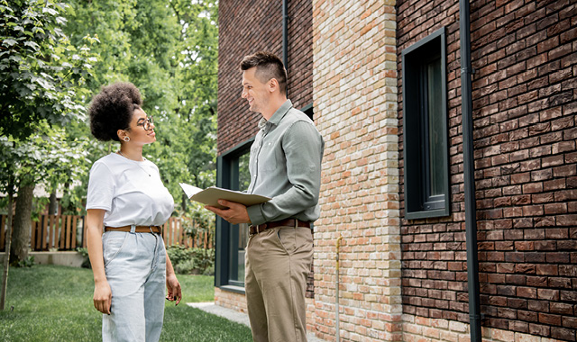 A realtor holds a folder and speaks to a woman outside a brick home in summer