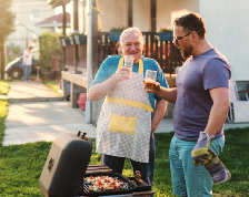 Elderly father and son enjoy glasses of iced tea while grilling in a backyard.