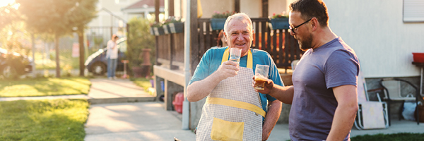 Elderly father and son enjoy glasses of iced tea while grilling in a backyard.