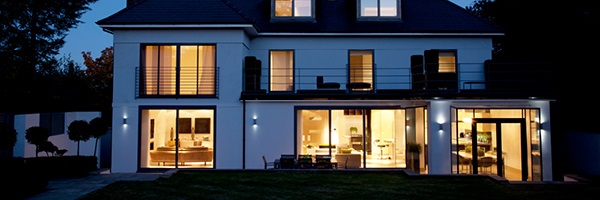 A white home with a dark roof is illuminated by indoor lights in several rooms at sunset.