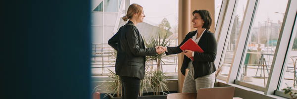 Two women in business attire in an office setting stand, shake hands, and smile at each other.