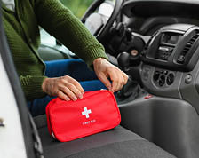 A driver in a parked vehicle reaches into a red first aid kit sitting on the passenger seat.