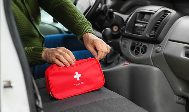 A driver in a parked vehicle reaches into a red first aid kit sitting on the passenger seat.