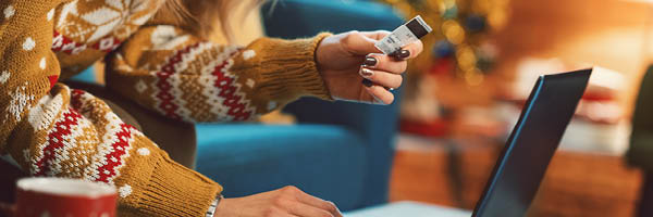 A woman sits on the couch in a cozy living room decorated for the holidays. She uses a laptop and credit card to shop online.