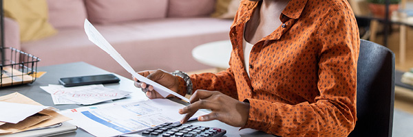 A woman with her hair in a bun sits at a table and works with papers and a calculator.