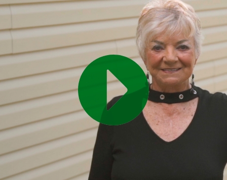 A senior woman stands in front of her home and smiles. A green play icon overlays the image.  