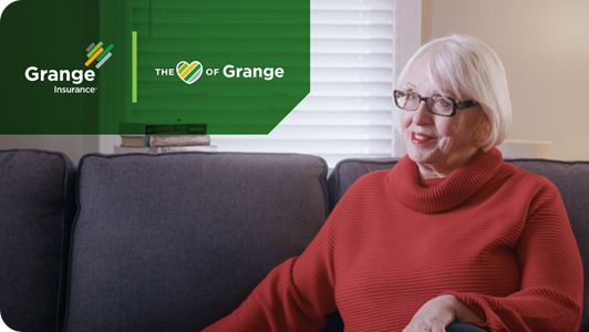 A senior woman wears a red sweater, sits on a couch and smiles while speaking.