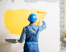 Man wearing a blue outfit paints a blank wall yellow with a roller brush.