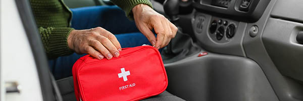 A driver in a parked vehicle reaches into a red first aid kit sitting on the passenger seat.